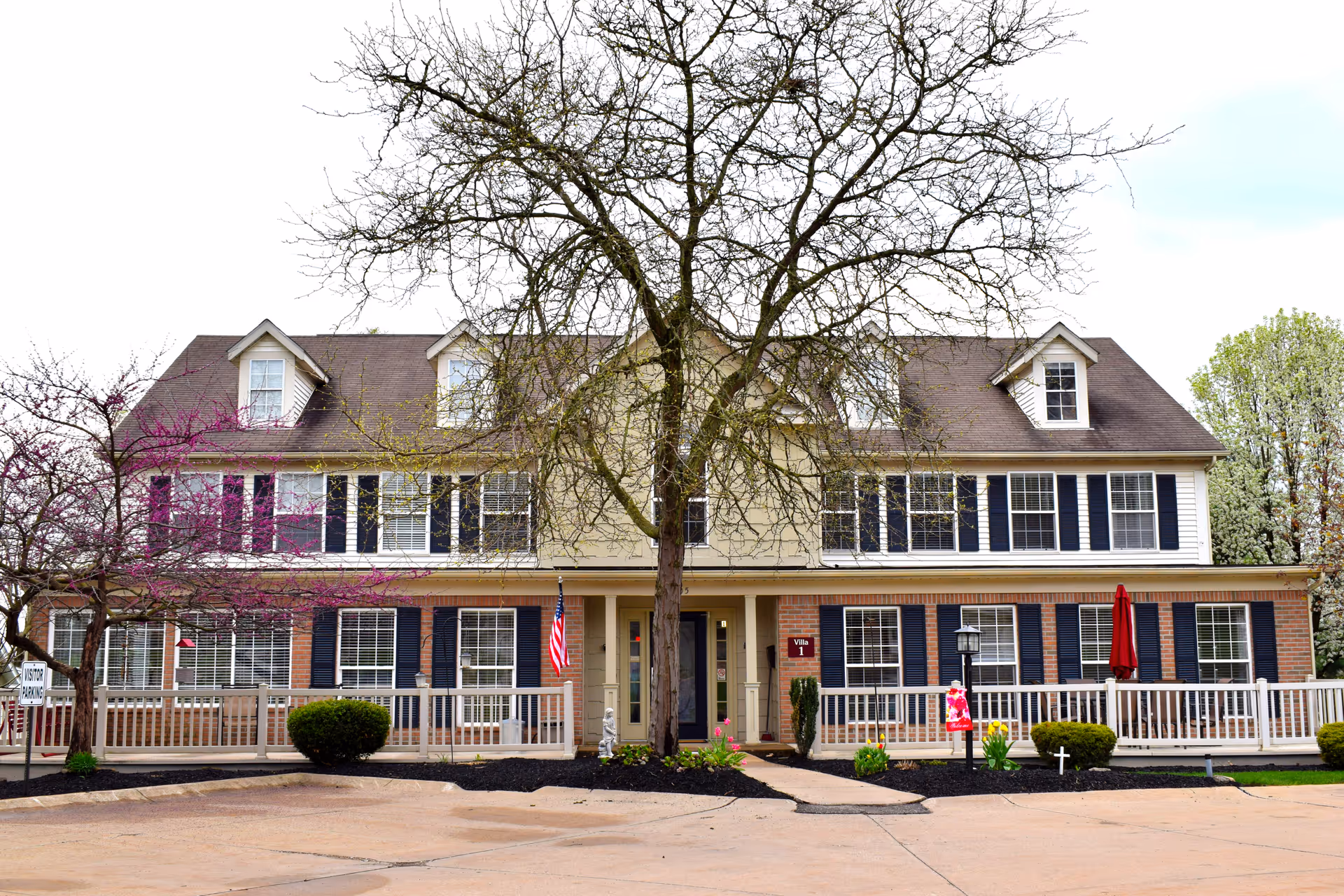 Front exterior view of a two-story assisted living facility building with multiple windows, black shutters, a central entrance, and a large tree in front. There are small bushes, flowers, and a flag near the entrance, with a paved driveway in the foreground.