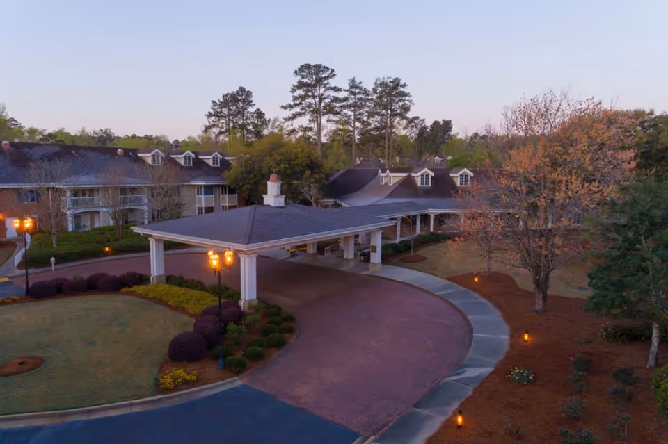 Exterior view of Brandon Wilde senior living facility at dusk, showing a covered driveway entrance with lit street lamps, landscaped bushes, trees, and a multi-story building with dormer windows in the background.