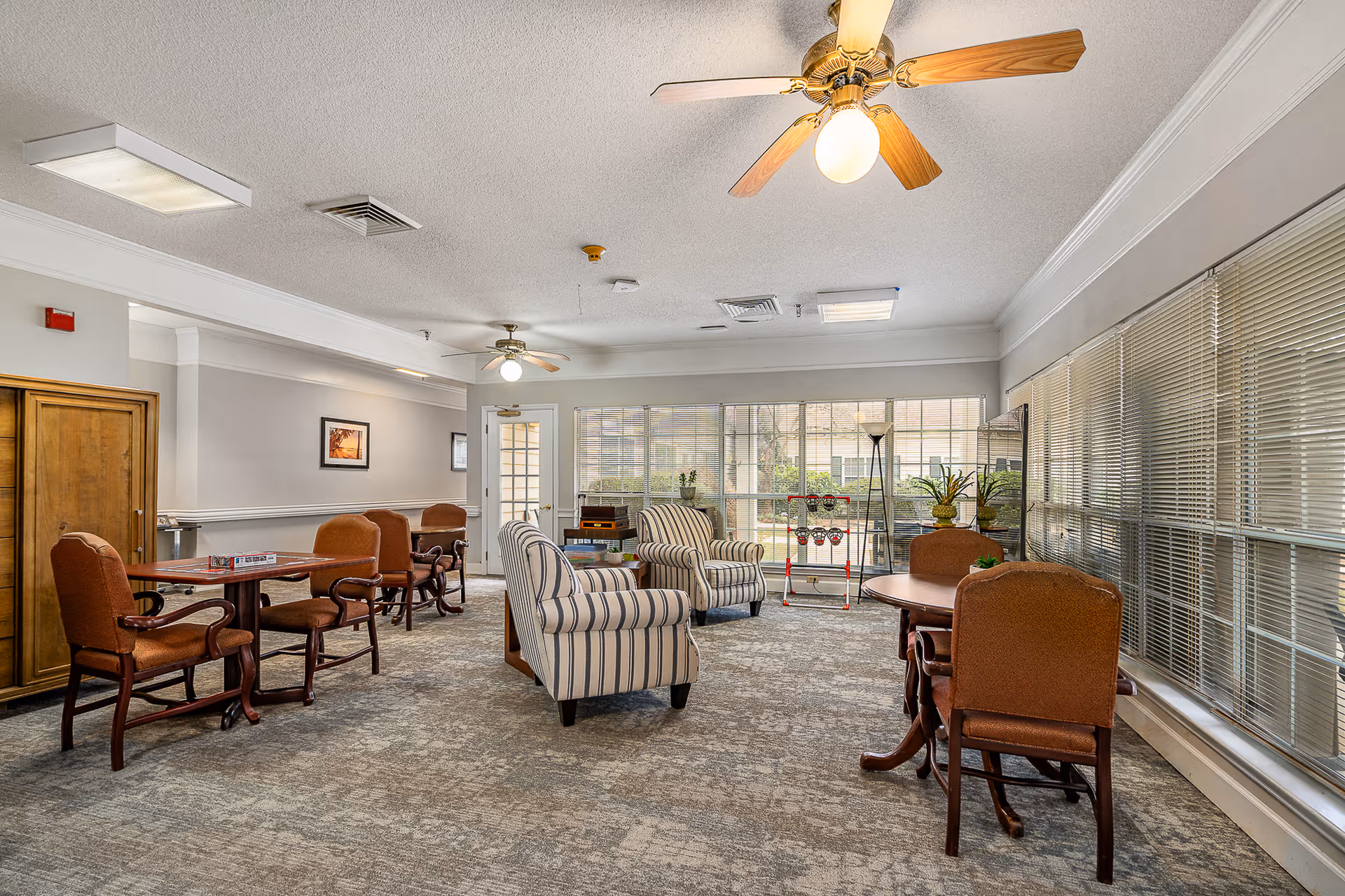 Sunlit senior living common room with upholstered armchairs, tables, and large windows overlooking a courtyard.