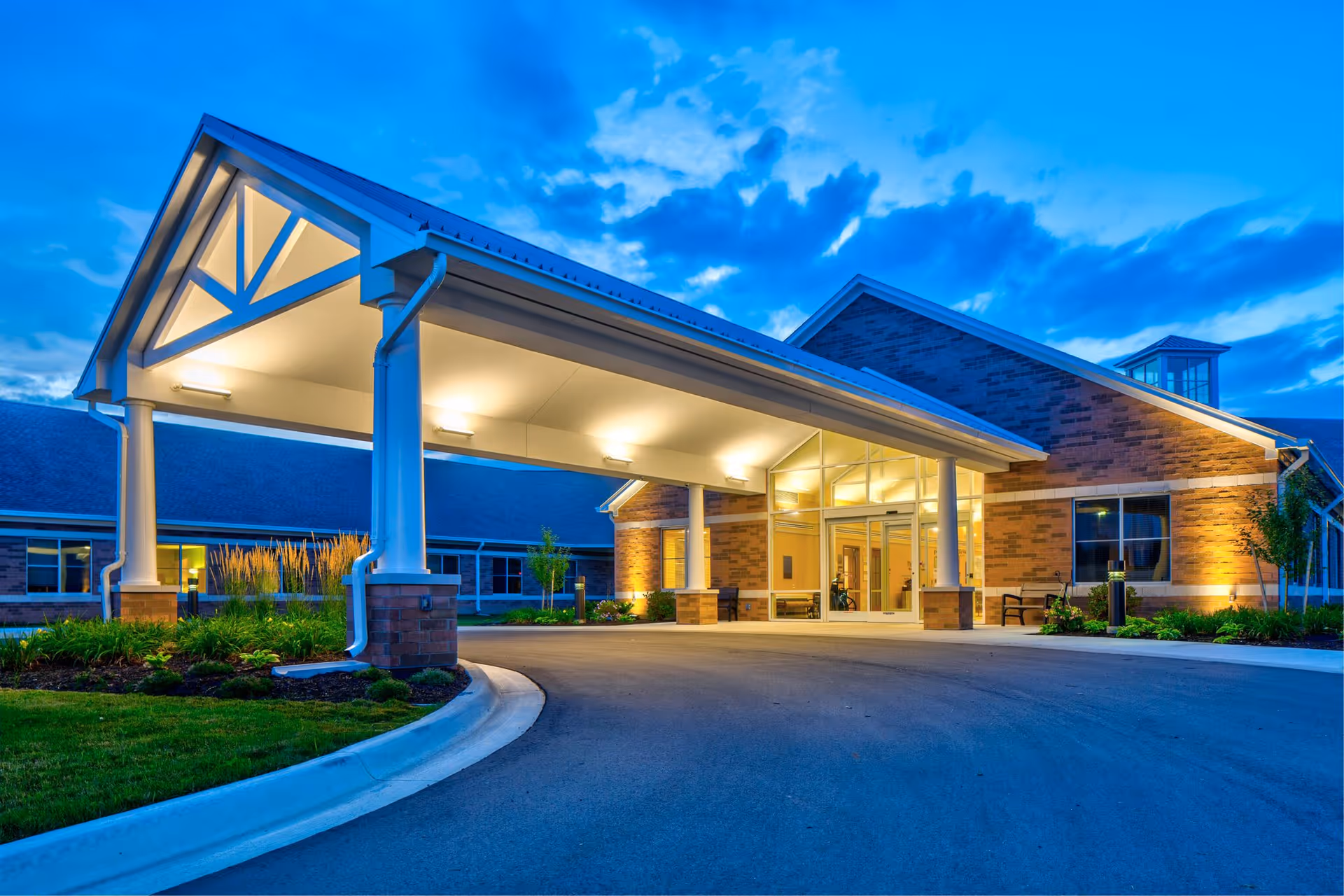 Exterior view of Pleasant View (Shiawassee County Medical Care Facility) at dusk, showing the building entrance with a covered drop-off area illuminated by warm lights, surrounded by landscaped greenery and a curved driveway.