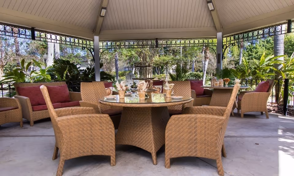 Outdoor covered seating area with a round glass-top table surrounded by wicker chairs and sofas with red cushions, set in a garden with lush greenery and palm trees visible in the background.