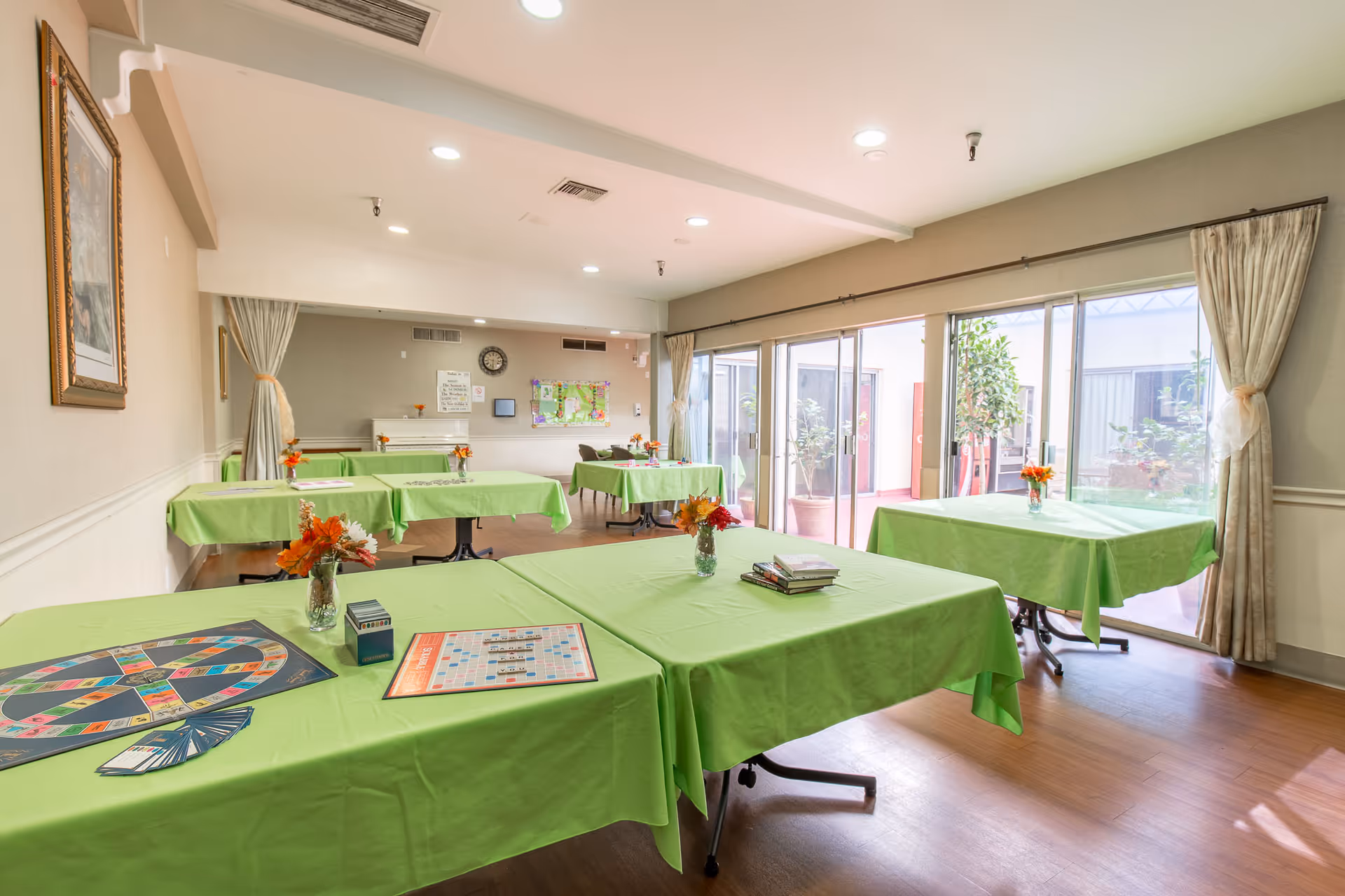 Bright activity/dining room with multiple tables covered in green tablecloths, game boards and small flower vases, and large windows letting in natural light.