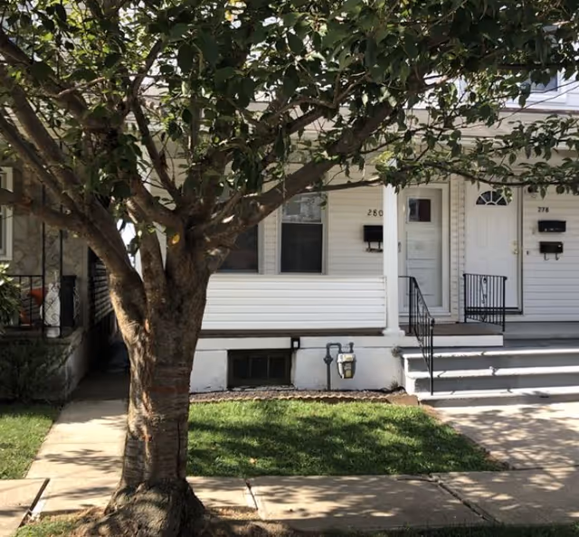 White residential-style facility front with doors, stairs, mailboxes, and a large tree.