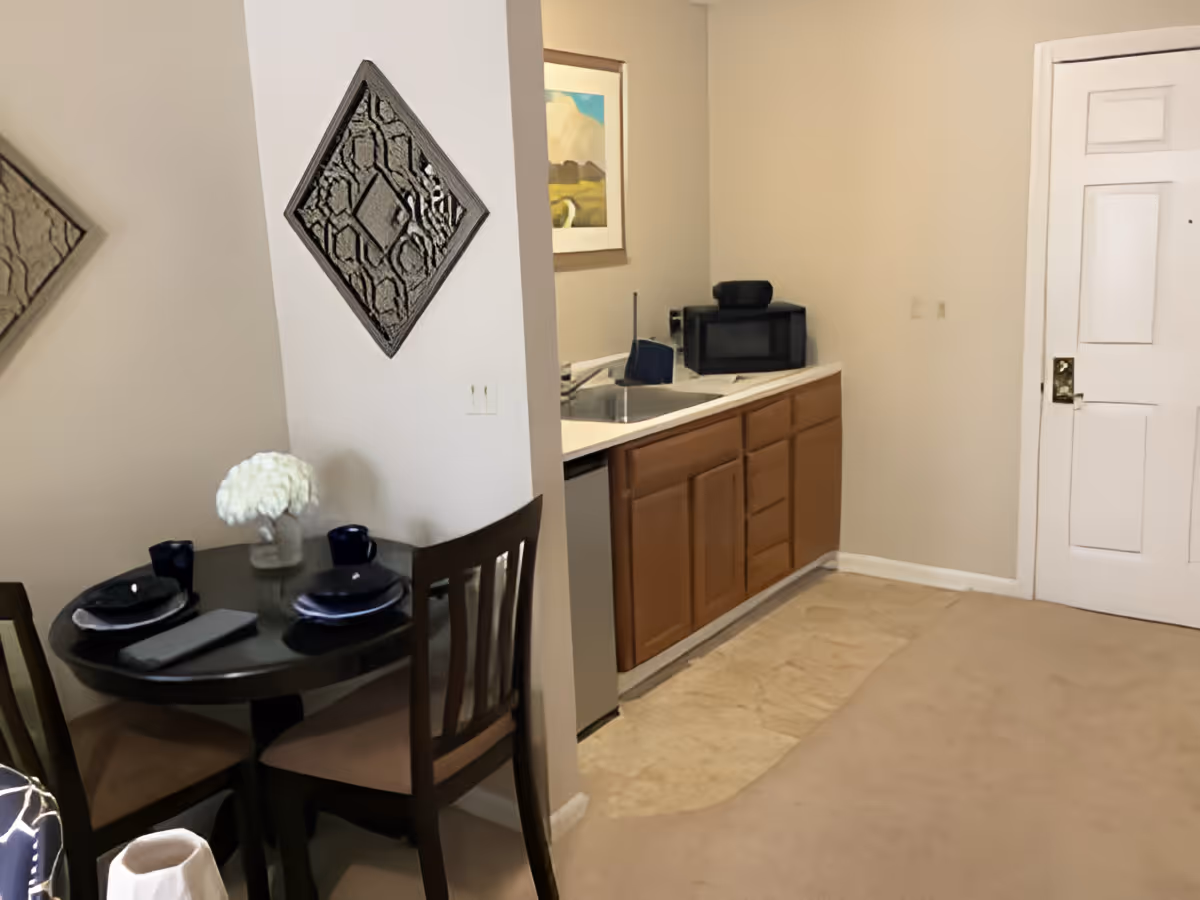 Interior view of a small dining and kitchen area in a senior living facility. The dining area has a round dark wooden table set with two place settings and a vase of white flowers. The kitchen area features a countertop with a sink, a microwave, and wooden cabinets below. A white door is visible in the background, and the walls are painted beige with a decorative wall hanging and framed artwork.
