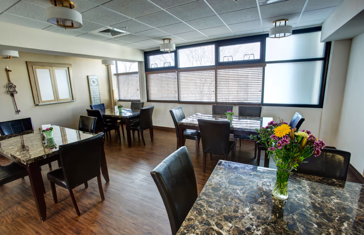 A bright dining room with several marble-patterned tables and black chairs arranged neatly. Each table has a small vase with colorful flowers. Large windows with blinds allow natural light to fill the room. The floor is wooden, and the ceiling has recessed lighting fixtures.