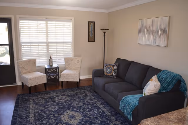 A cozy living room with a dark gray sofa adorned with two decorative pillows and a teal throw blanket. Two beige patterned chairs are placed near a window with white blinds, separated by a small dark wooden table holding a decorative item. A tall floor lamp stands in the corner, and a framed abstract painting hangs on the beige wall above the sofa. The floor is covered with a large blue patterned rug.