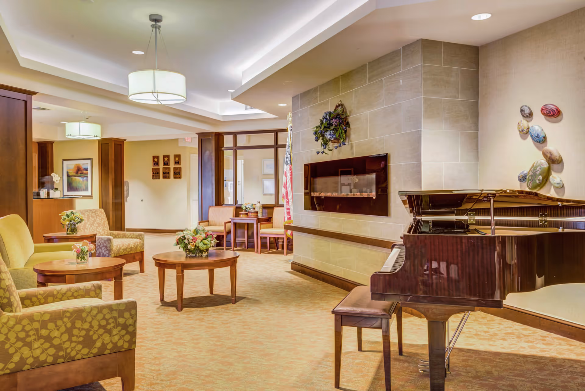 A cozy senior living facility common area with comfortable armchairs and sofas arranged around wooden coffee tables. A grand piano is positioned near a modern wall-mounted fireplace with a decorative wreath above it. The room is warmly lit with ceiling lights and decorated with wall art and floral arrangements.