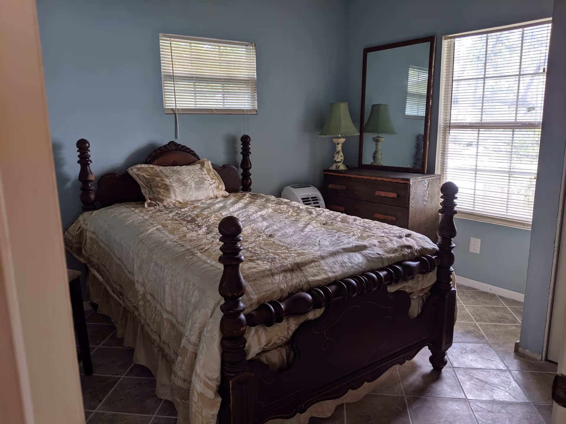 A small bedroom with a wooden bed frame and a beige patterned comforter. There is a wooden dresser with a large mirror and a green lamp on top. Two windows with blinds allow natural light into the room. The walls are painted light blue and the floor is tiled.