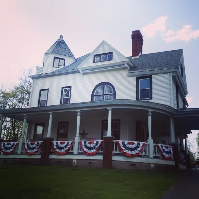 Large white Victorian-style house with a wraparound porch decorated with red, white, and blue bunting.