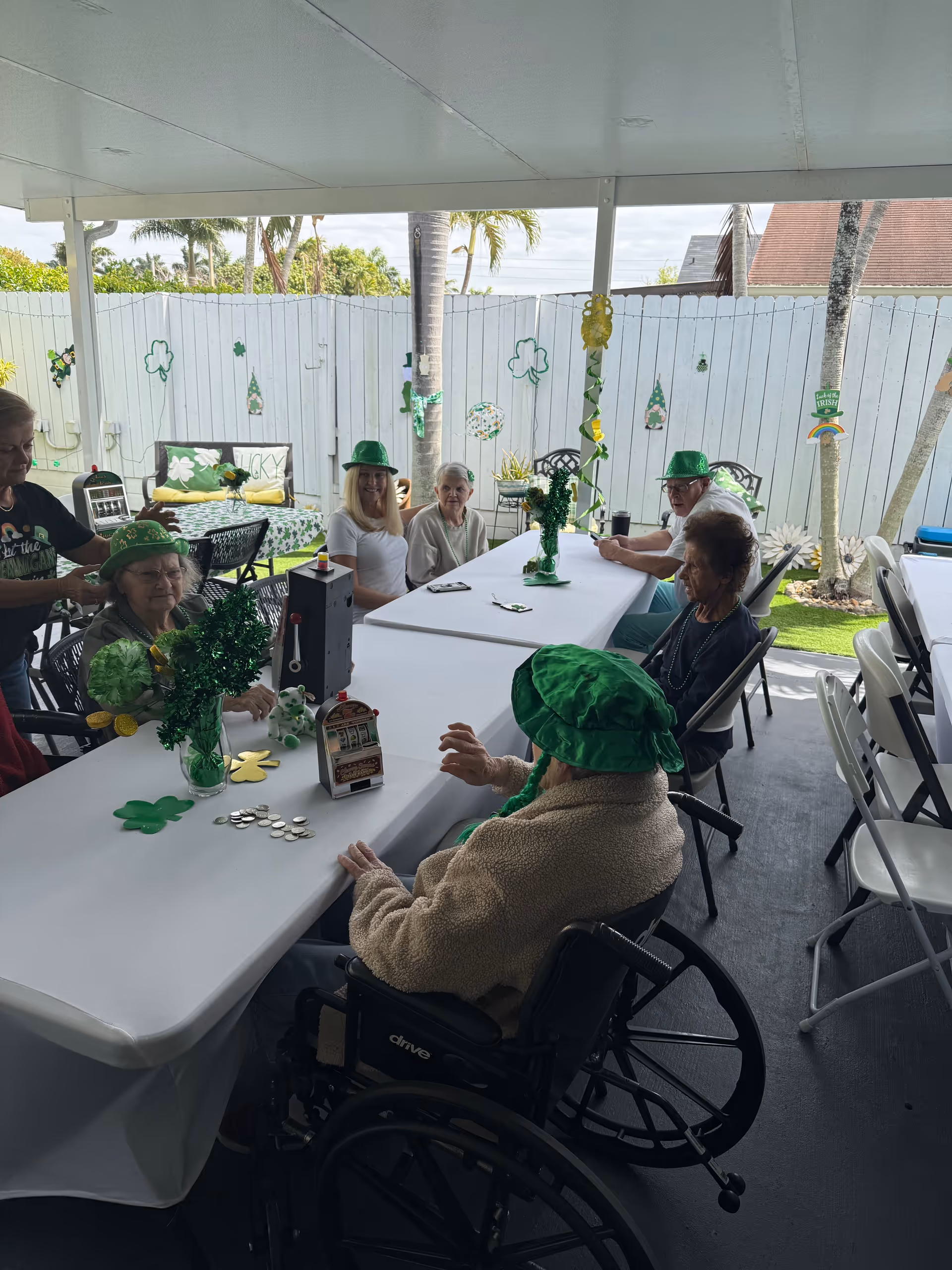 Several elderly residents wearing green hats sit around a long decorated table under a covered outdoor patio.