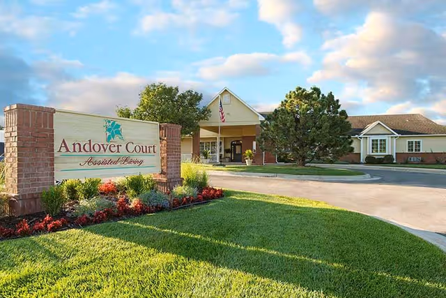 Exterior view of Andover Court Assisted Living facility with a large sign in the foreground surrounded by landscaped flowers and greenery. The building is visible in the background under a partly cloudy sky.