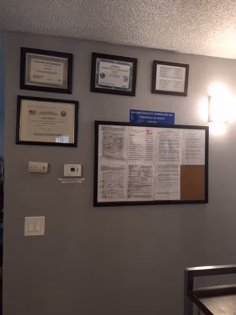 Interior wall of an office with framed certificates and documents displayed. There is a thermostat and light switches on the wall, and a light fixture illuminating the area. A sign reads 'Solano Quality Homecare Inc Fairfield CA 94533 Office'.