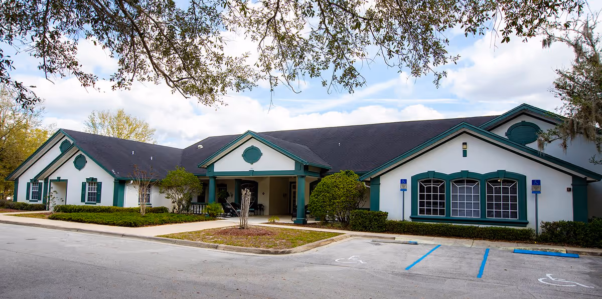 Exterior view of a single-story senior living facility building with white walls and green trim. The building has a dark roof and several large windows. There are two handicapped parking spaces in front, and trees with branches partially frame the top of the image.