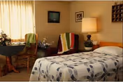 A cozy bedroom in a nursing and rehab facility featuring a bed with a patterned quilt, a wooden chair with a colorful knitted blanket draped over it, a small round table with a flower arrangement, a lamp on a nightstand, and framed pictures on the beige walls.