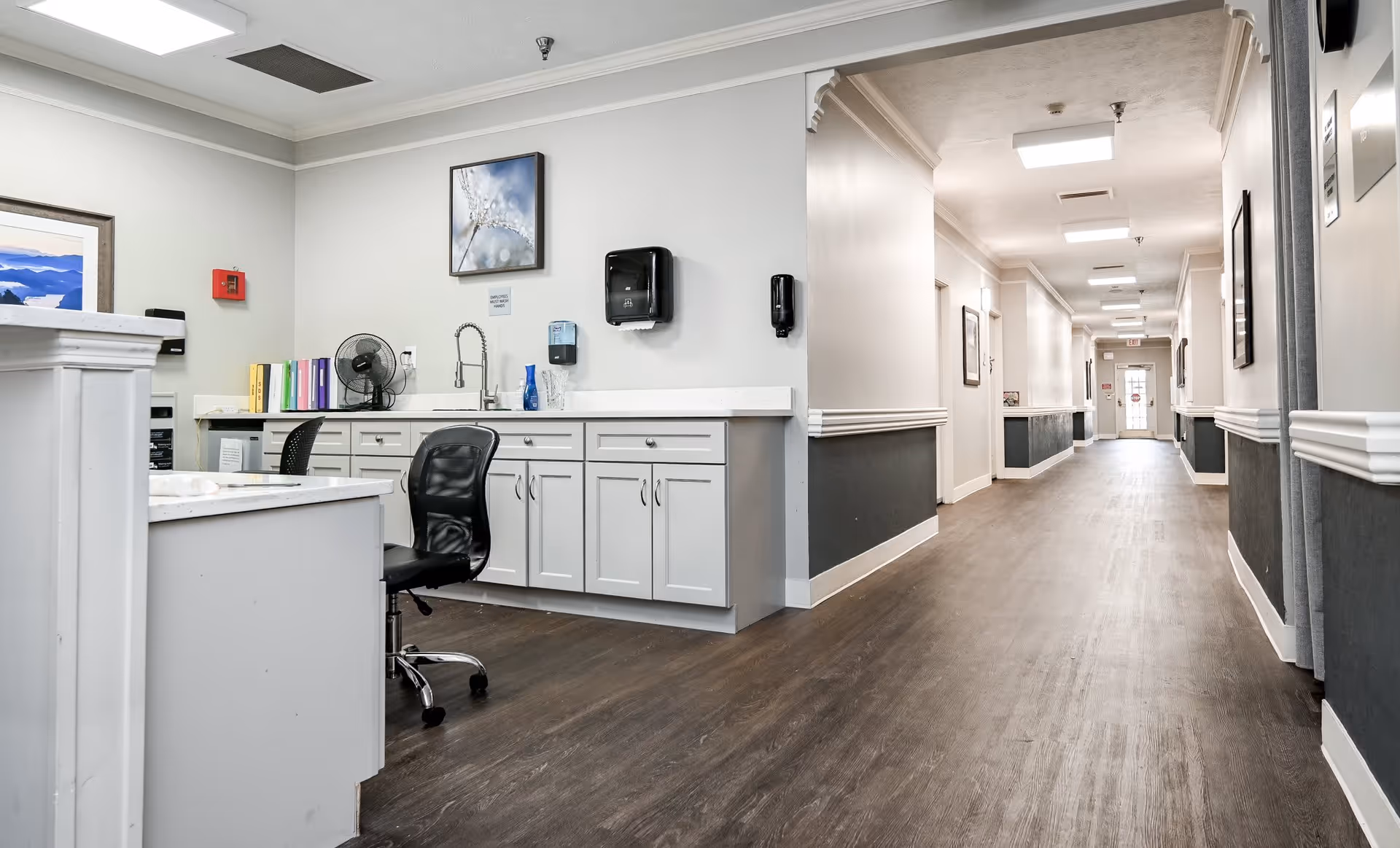 Interior view of a senior living facility hallway with a reception desk area on the left. The desk has a black office chair, a fan, colorful binders, a sink, and wall-mounted dispensers. The hallway extends to a door with exit signs and framed pictures on the walls.