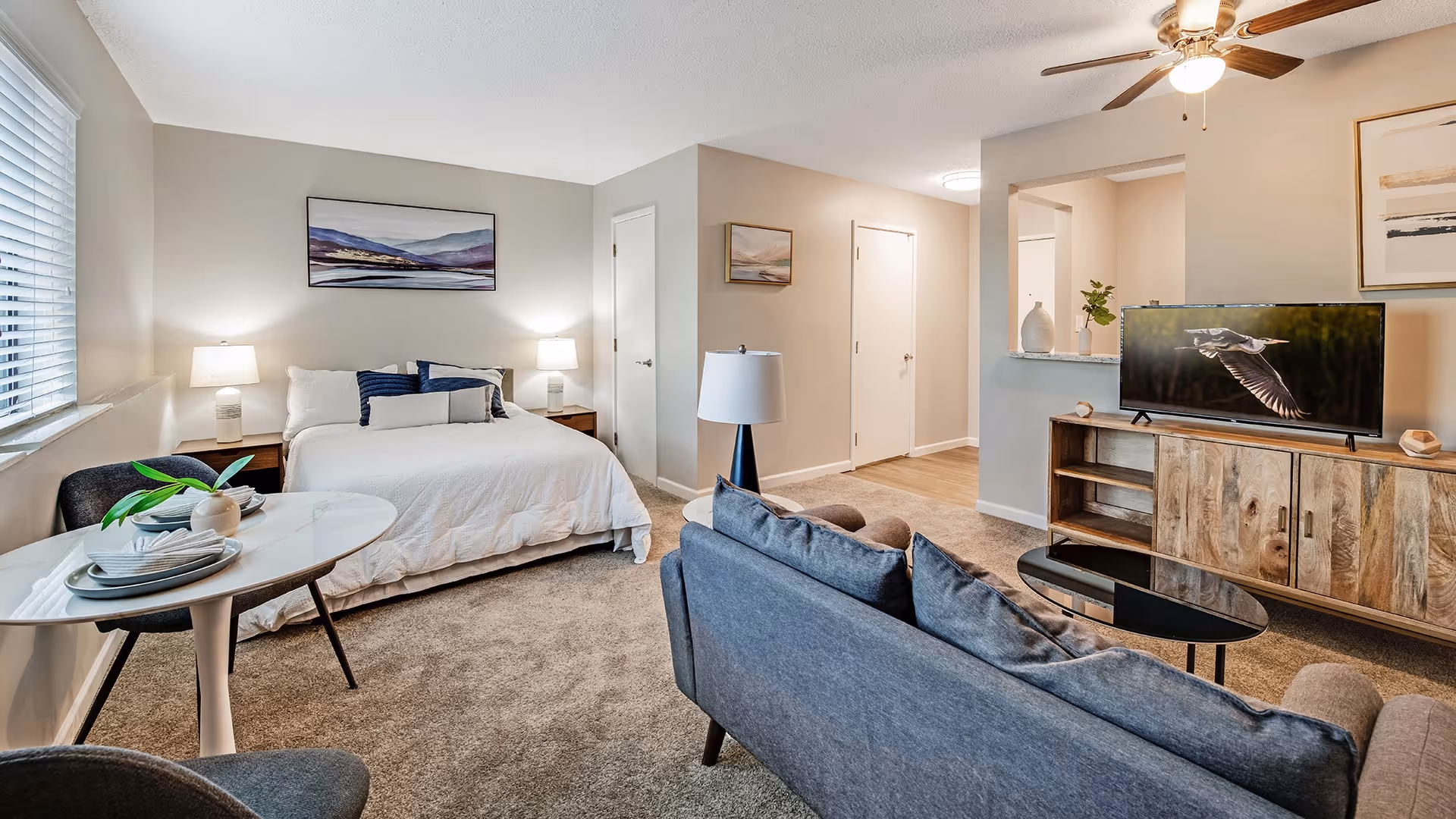 A bright and cozy studio apartment featuring a bed with white bedding and blue accent pillows, two bedside tables with lamps, a small round dining table set for two with plates and napkins, a gray sofa facing a wooden TV stand with a flat-screen TV displaying a bird in flight, and neutral-colored walls with framed artwork. The room has a window with blinds and a ceiling fan with a light.