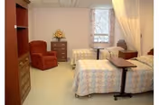 A shared senior bedroom with two twin beds, a recliner, dressers, overbed table and a privacy curtain.