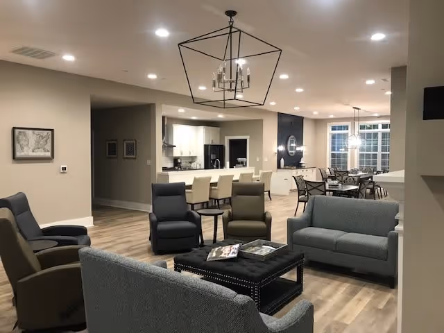 Open communal living area with sofas, armchairs and a central ottoman, overlooking a kitchen with bar seating and a dining table.