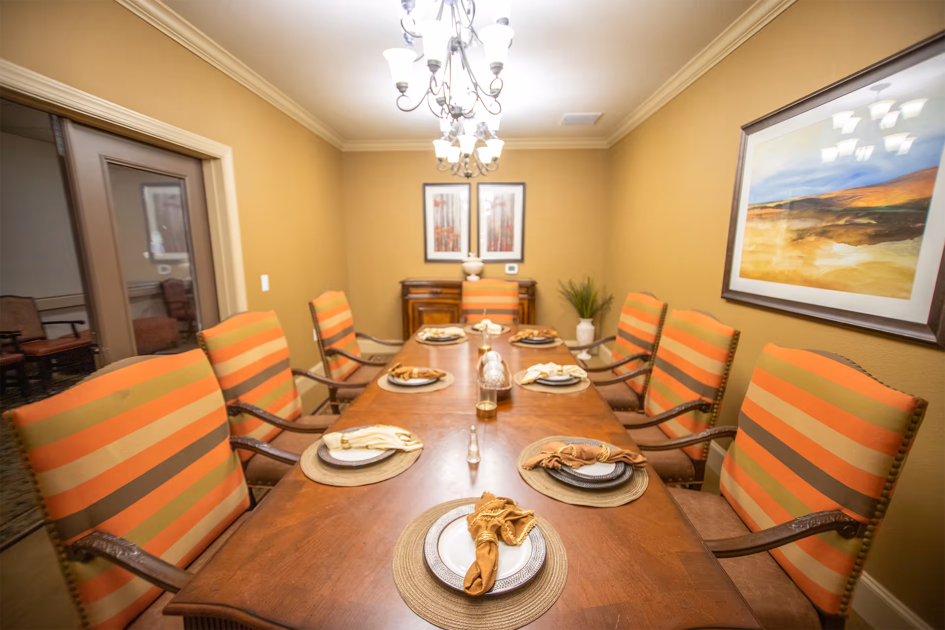 A dining room with a long wooden table set for six people. The chairs have orange and brown striped upholstery. The table is set with plates, napkins, and placemats. There are two framed pictures on the far wall, a chandelier hanging from the ceiling, and a large framed painting on the right wall. A plant is placed in the corner near the back wall.