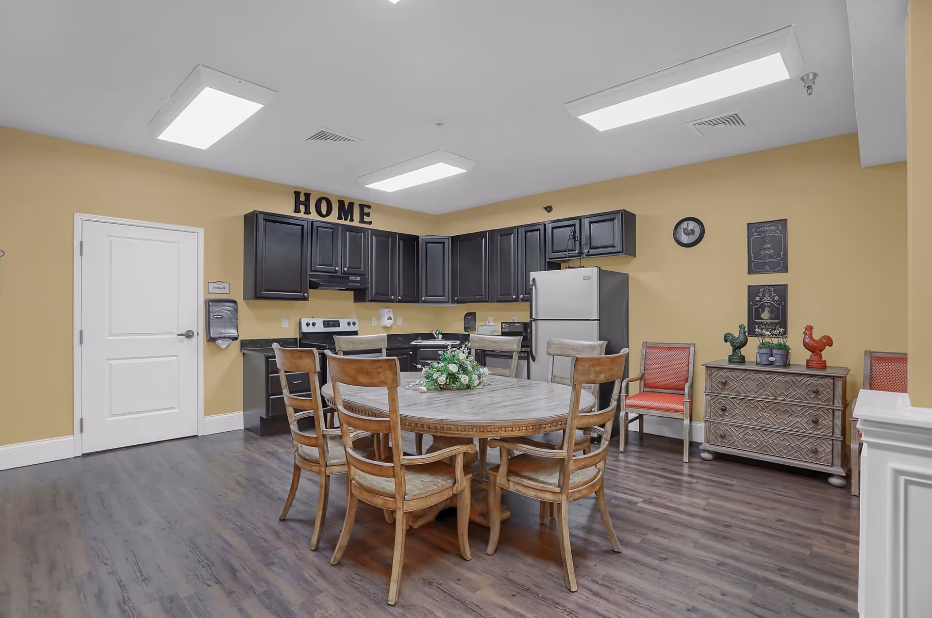 Communal dining area with a round wooden table and chairs in front of a kitchenette with black cabinets and a refrigerator, with 'HOME' letters displayed above the cabinets.