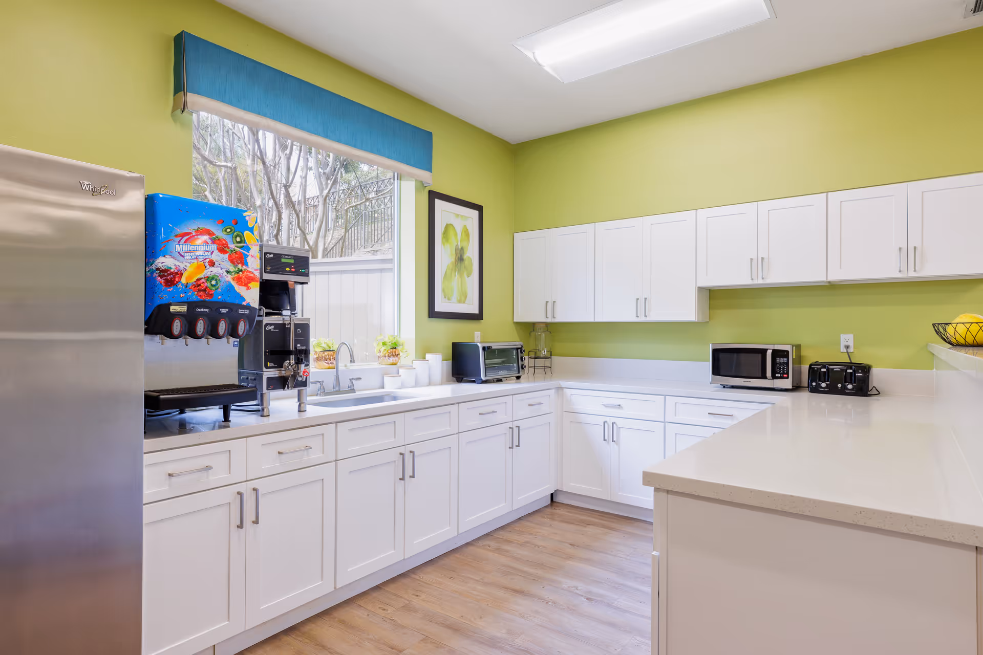 Bright kitchen with white cabinets and countertops, a stainless steel refrigerator, a beverage dispenser, a toaster oven, a microwave, a toaster, and a window with a blue valance overlooking trees outside. The walls are painted light green and the floor is wood.