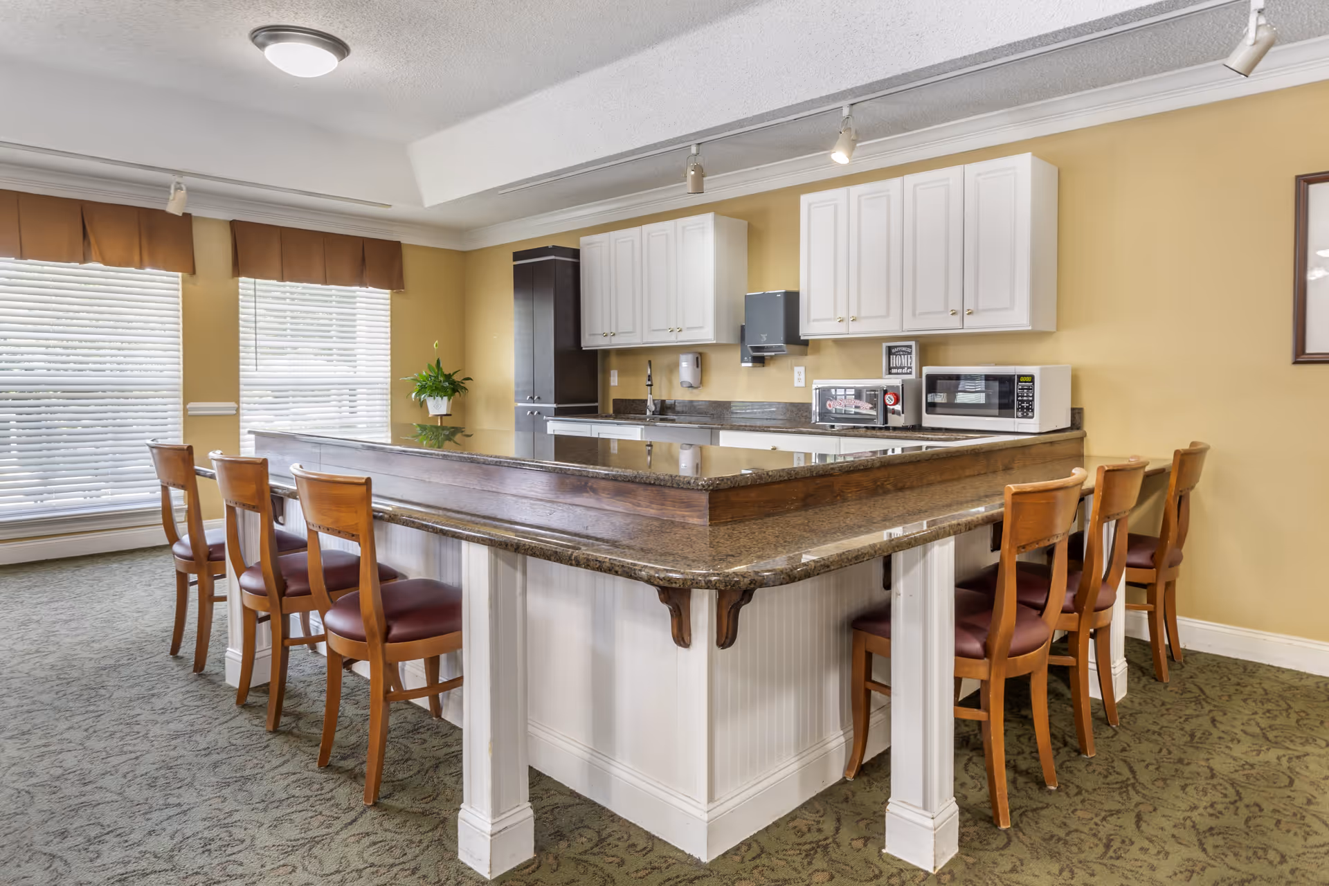 A kitchen area with a large granite countertop island surrounded by wooden chairs with red cushions. The kitchen has white cabinets, a microwave, a toaster oven, and a paper towel dispenser mounted on a yellow wall. Two large windows with white blinds and brown valances allow natural light into the room. The floor is carpeted with a patterned green carpet.