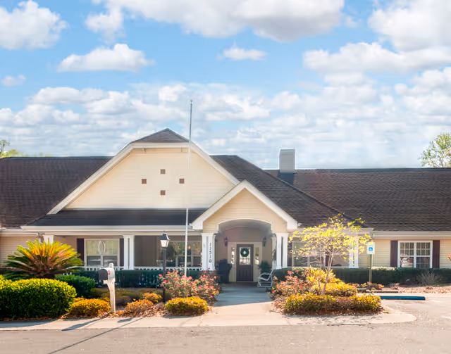 Front entrance of a single-story senior living facility with a covered porch, flagpole, and landscaped beds under a blue sky.