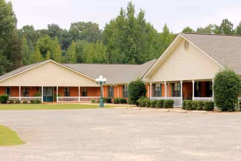 A single-story senior living facility with beige siding, a wide front porch, and surrounding trees.