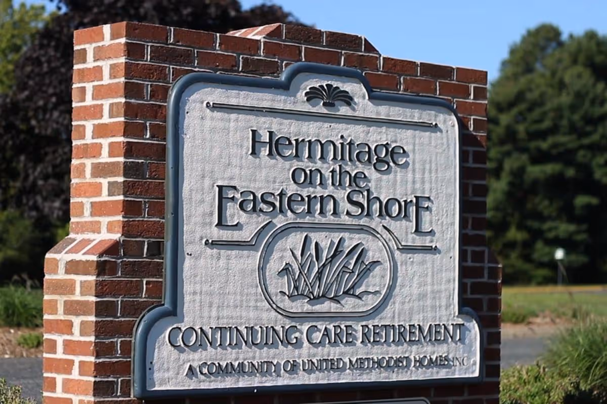 A brick signpost with a white and blue plaque that reads 'Hermitage on the Eastern Shore Continuing Care Retirement A Community of United Methodist Homes Inc' with greenery and trees in the background under a clear blue sky.