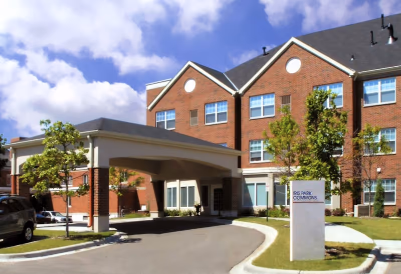 Exterior view of a three-story brick assisted living facility with a covered entrance driveway, several windows, small trees, and a sign that reads Iris Park Commons.