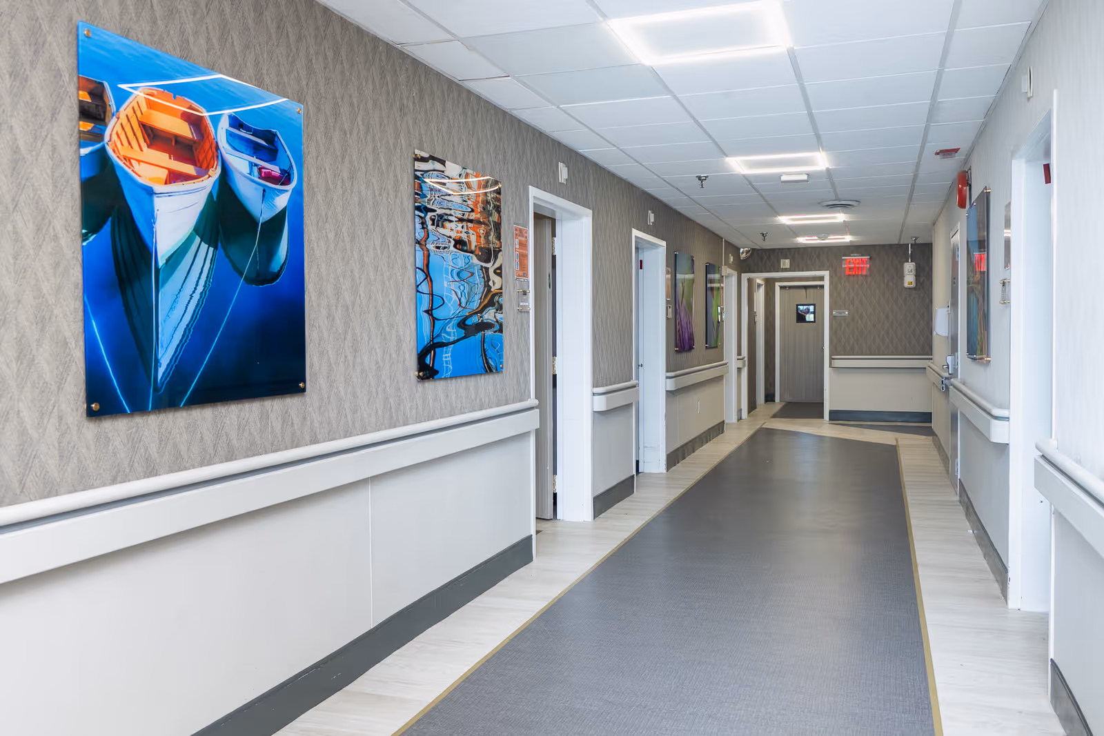 A well-lit hallway in a rehabilitation and nursing center with light-colored walls, handrails on both sides, several closed doors, and colorful artwork featuring boats and abstract reflections hanging on the walls.