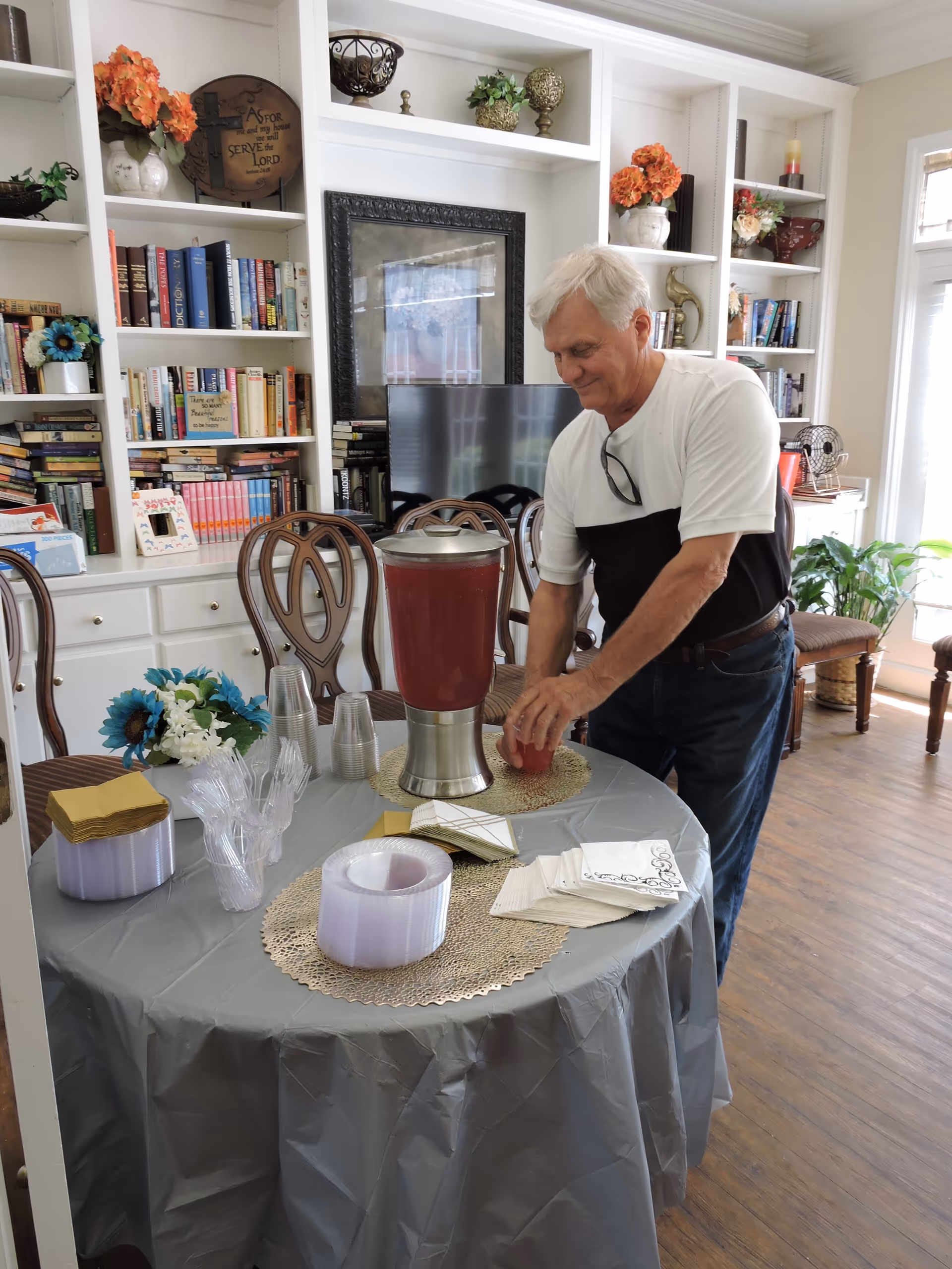 A man arranges cups and napkins at a round table with a drink dispenser in a furnished senior living common room with bookshelves and chairs.
