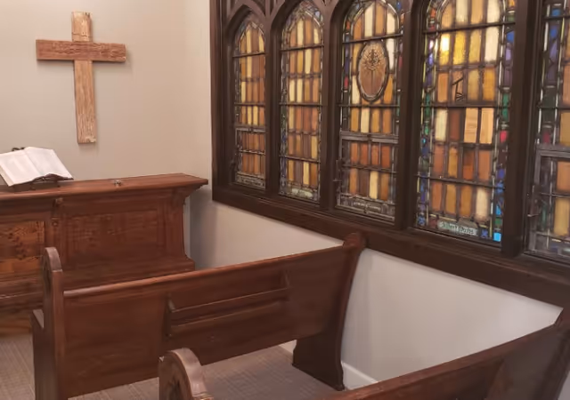 Small chapel room with wooden pews, a wooden altar holding an open book, a wooden cross mounted on the wall, and stained glass windows with colorful patterns.
