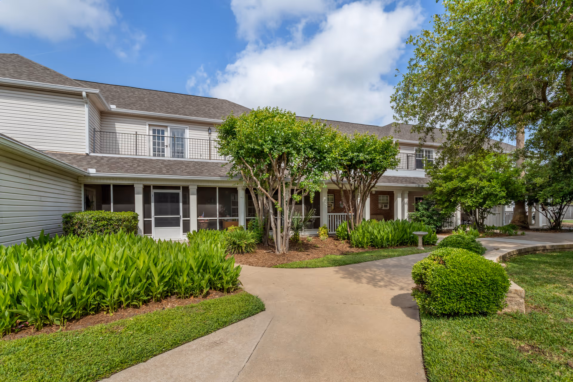 Front exterior of a two-story senior living building with a paved walkway, landscaped gardens, screened porches and trees under a partly cloudy sky.