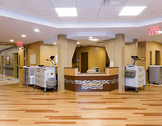 Interior view of a rehabilitation and nursing center reception area with a central curved desk, medical carts on either side, beige walls, wood-patterned flooring, and ceiling lights. Exit signs are visible above doorways in the background.