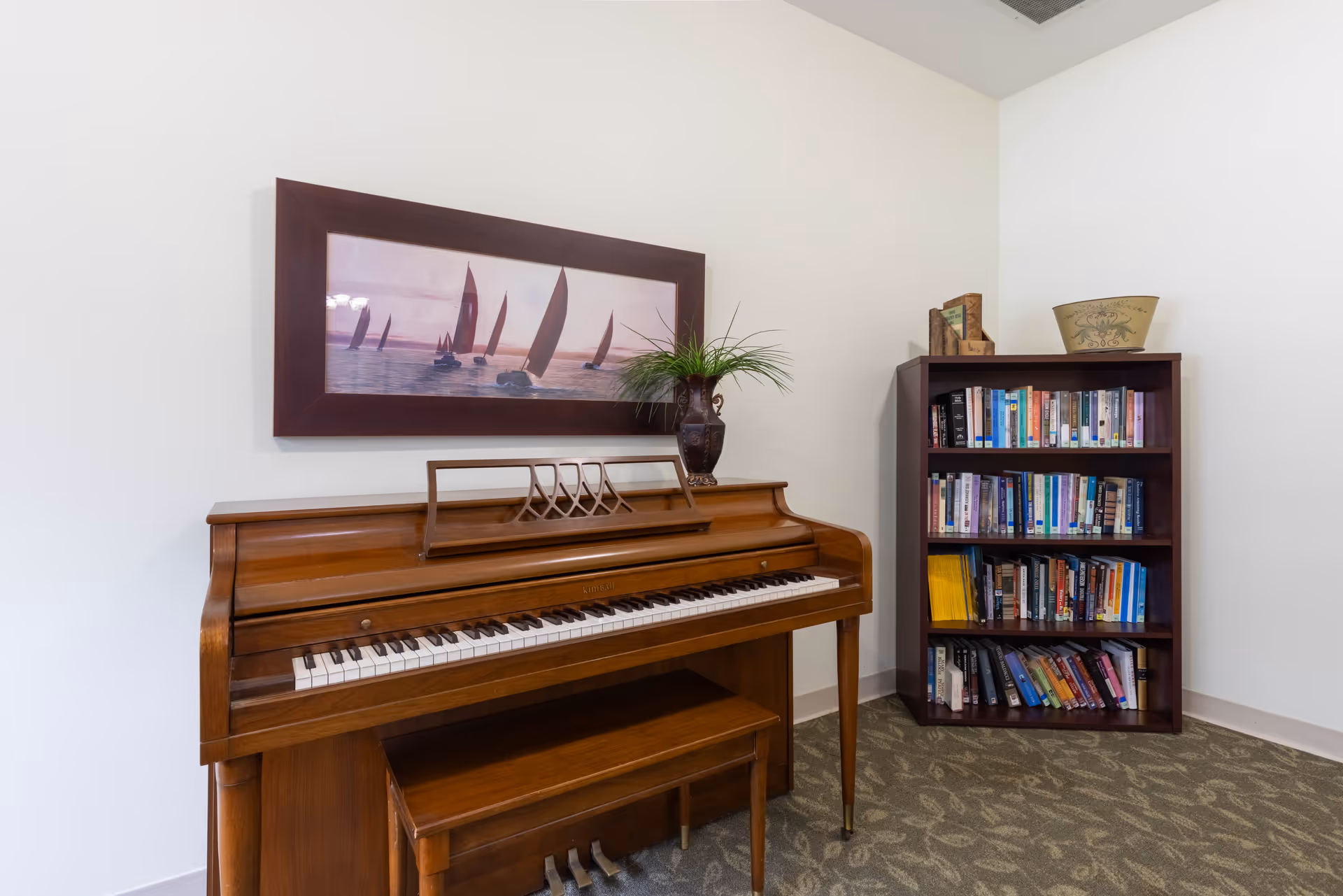 Wooden upright piano with a bench beneath a framed sailboat painting, next to a bookshelf filled with books and a decorative vase.