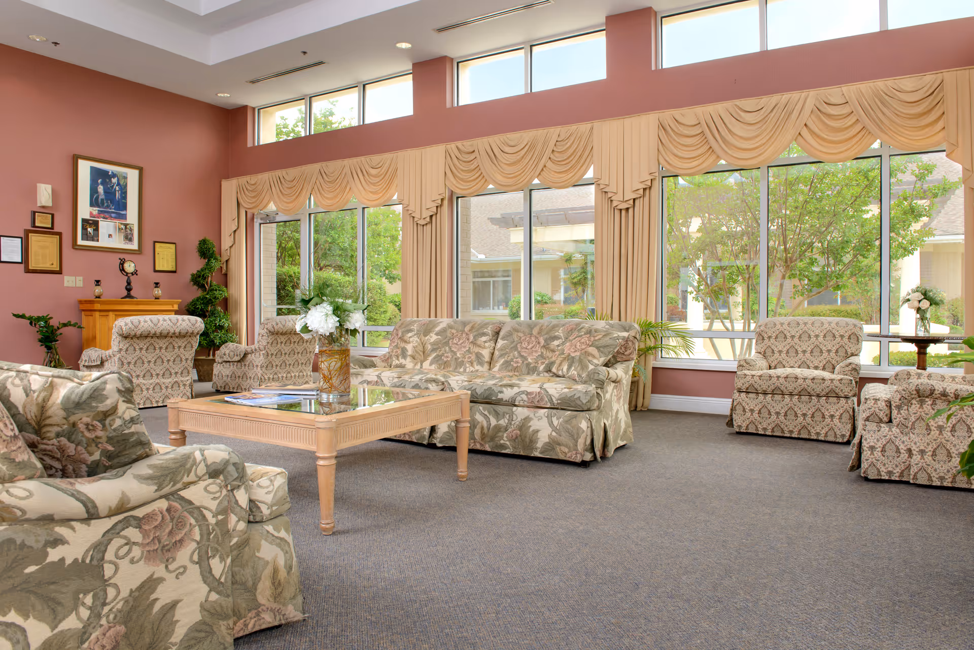 A bright and spacious living room area with large windows letting in natural light. The room features floral patterned sofas and armchairs arranged around a wooden coffee table with a vase of white flowers. The walls are painted a warm mauve color, and there are framed pictures and certificates on one wall. Outside the windows, green trees and parts of the building exterior are visible.