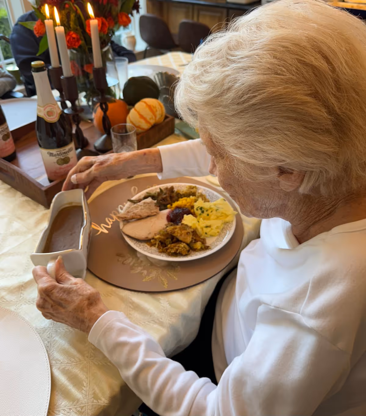 An elderly woman with white hair is seated at a dining table, holding a gravy boat and pouring gravy onto a plate filled with a variety of food including turkey, mashed potatoes, stuffing, and cranberry sauce. The table is decorated with candles, small pumpkins, and bottles of sparkling beverages.