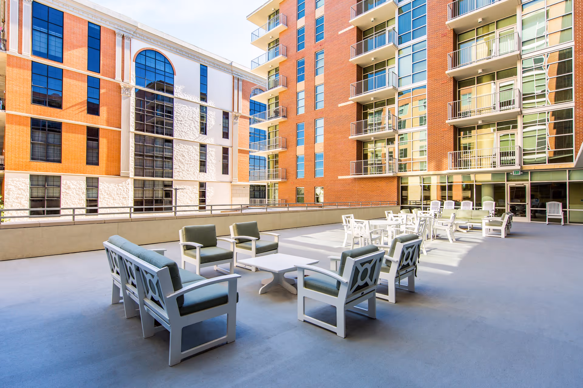 Outdoor patio area at The Village of River Oaks featuring multiple seating arrangements with cushioned chairs and tables, surrounded by multi-story brick and glass buildings under a clear sky.