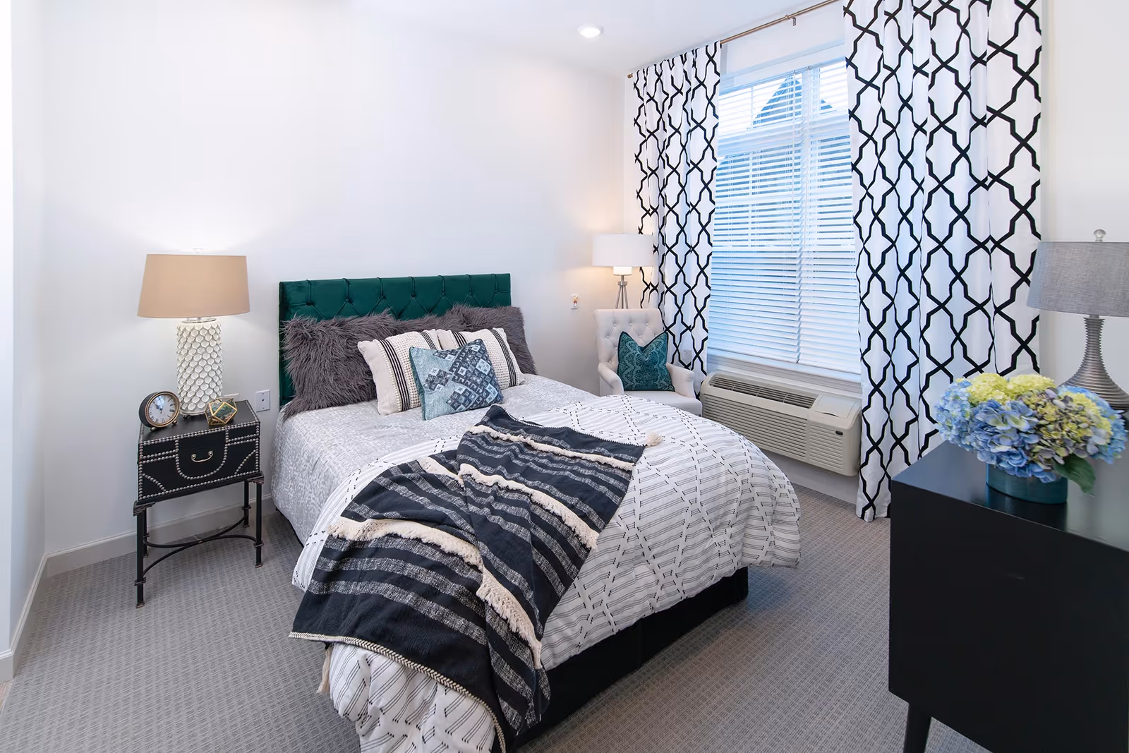 A neatly made bed with a green tufted headboard, multiple decorative pillows, and a black and white striped throw blanket in a bright bedroom. The room has patterned black and white curtains, a window with blinds, a beige armchair with a green pillow, two bedside lamps, a small black nightstand with a clock and decorative item, and a black dresser with a vase of blue and green flowers.