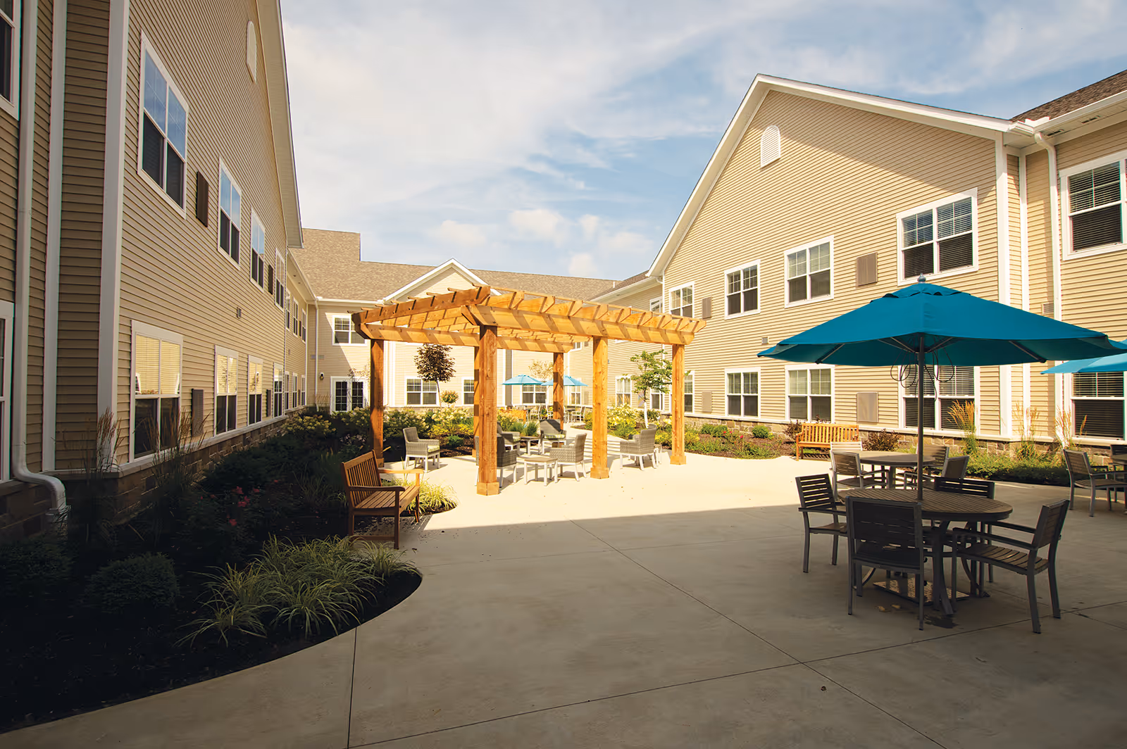 Outdoor courtyard area of a senior living facility with beige two-story buildings surrounding a concrete patio. The patio features a wooden pergola with seating underneath, several tables with blue umbrellas, chairs, benches, and landscaped garden beds along the edges.