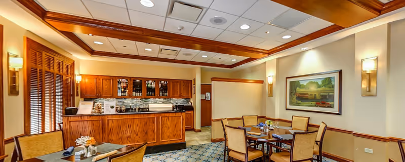 A cozy dining area in Westminster Place featuring wooden tables and chairs with place settings including napkins and small flower arrangements. The room has warm lighting with wall sconces, a wooden ceiling trim, and a framed painting on the wall. There is a wooden cabinet and counter area in the background with glass-fronted upper cabinets and a tiled backsplash.