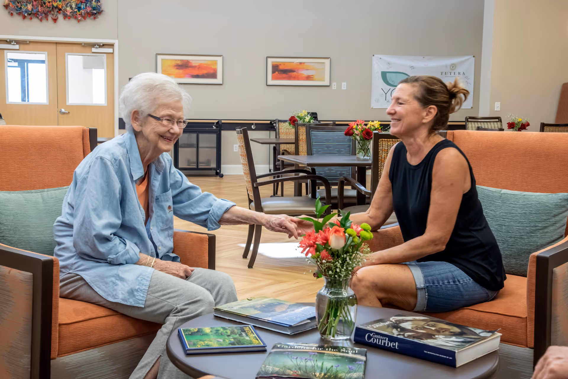 An elderly woman and a middle-aged woman sitting on orange armchairs in a bright, welcoming common area. They are smiling and holding hands across a round table with books and a vase of flowers. The background shows tables and chairs, colorful artwork on the walls, and large windows letting in natural light.
