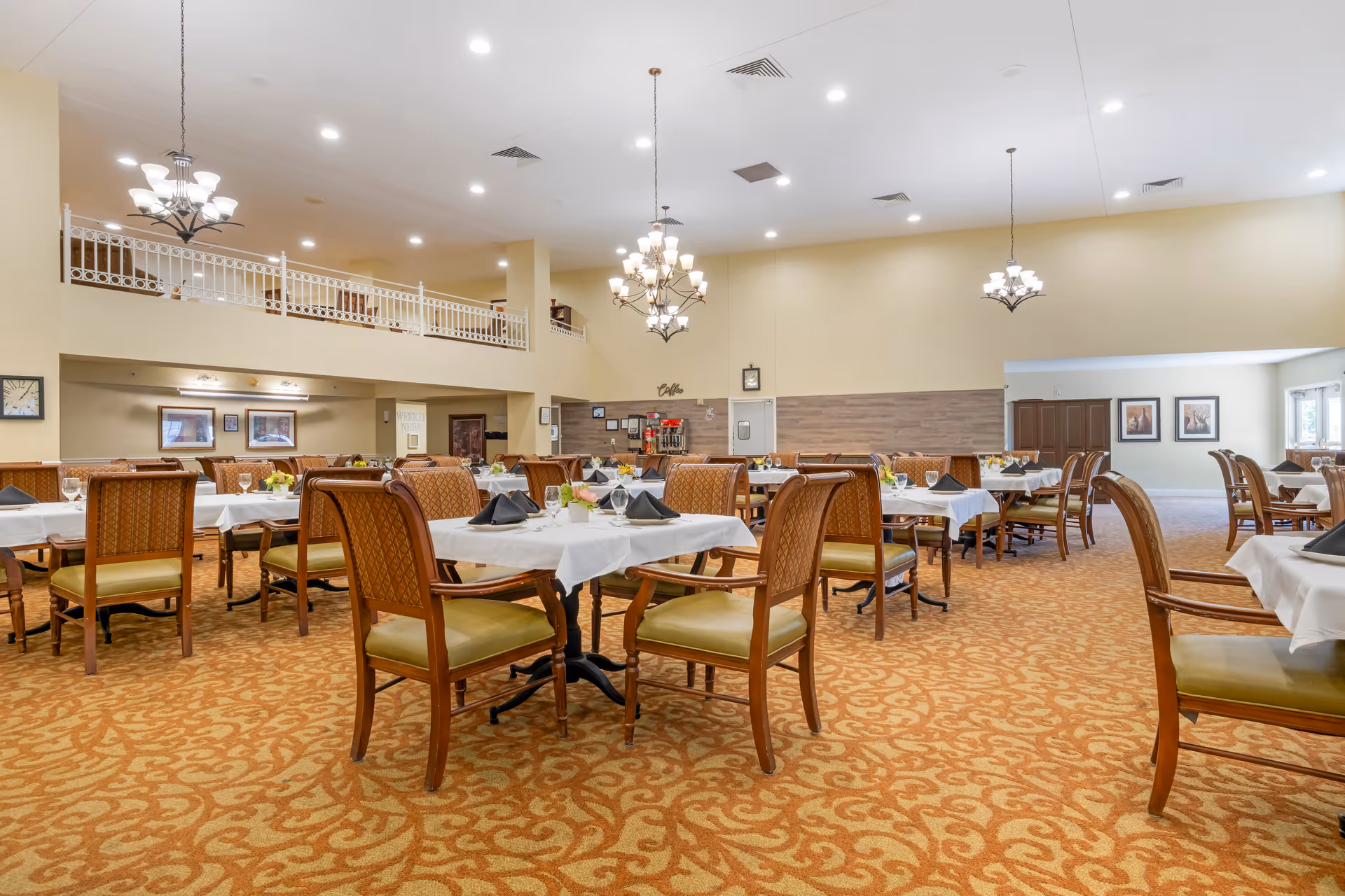 Spacious dining room with multiple tables covered in white tablecloths, each set with black napkins, glasses, and small floral centerpieces. The room features patterned carpet, wooden chairs with cushioned seats, and elegant chandeliers hanging from a high ceiling. There is a coffee station against the far wall and framed artwork on the walls.