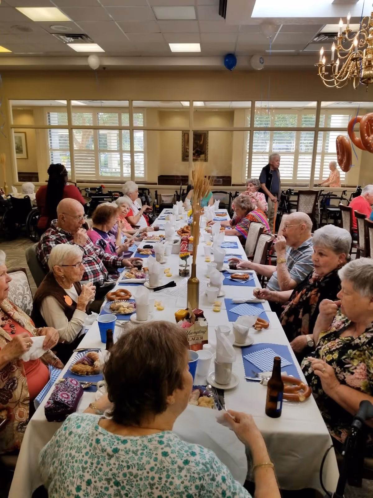 A group of elderly people seated around a long dining table in a well-lit room with large windows. The table is set with plates of food, pretzels, cups, and napkins. Some people are eating while others are engaged in conversation. The room has beige walls, a chandelier, and balloons tied to chairs.