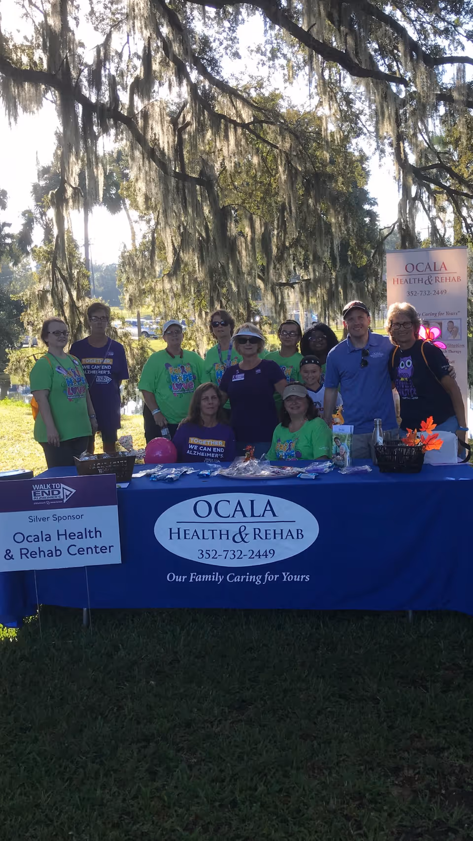 A group of people standing and sitting behind a table covered with a blue Ocala Health & Rehab Center tablecloth at an outdoor event. The table has various items on it, and there is a sign indicating Ocala Health & Rehab Center as a Silver Sponsor for the Walk to End Alzheimer's. The background shows trees with hanging moss and a bright sunny day.
