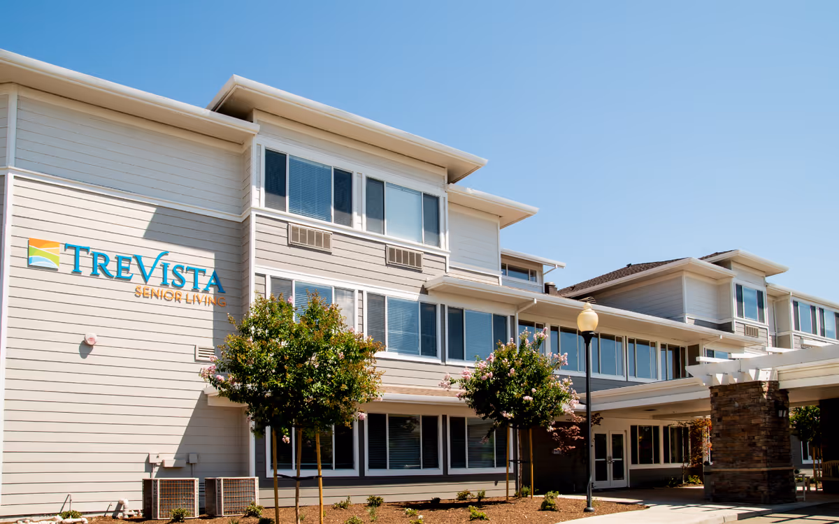 Exterior view of TreVista Senior Living facility showing a multi-story building with large windows, a covered entrance supported by stone pillars, small trees, and a clear blue sky.