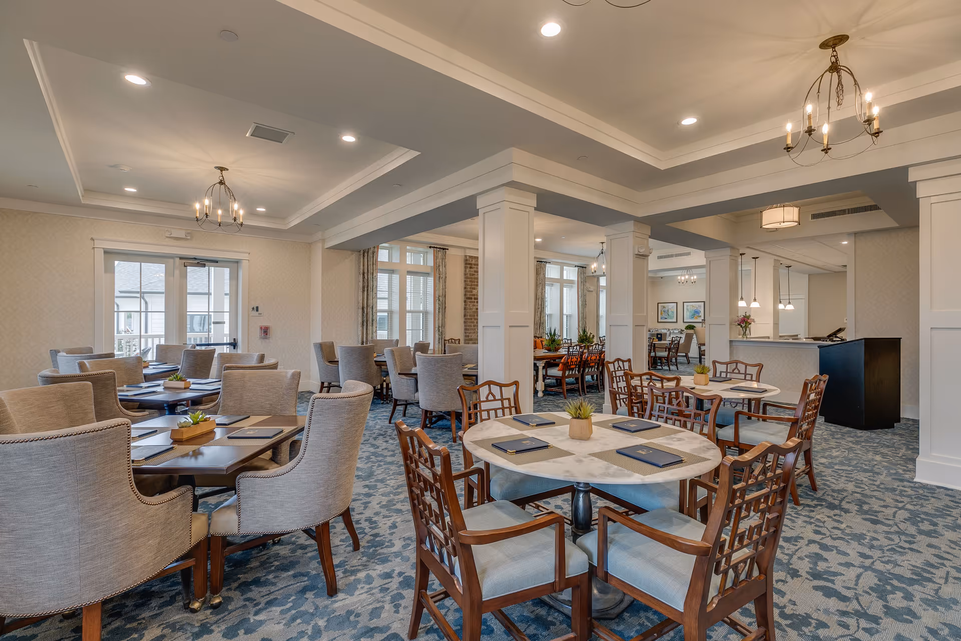 A spacious and well-lit dining room with multiple tables and chairs arranged neatly. The room features a patterned blue carpet, light-colored walls, and large windows with curtains allowing natural light to enter. There are chandeliers and recessed lighting on the ceiling, and small plants are placed on the tables for decoration.