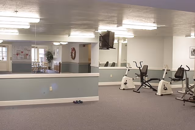 Interior exercise room at a retirement residence with stationary bikes, a wall-mounted TV, mirrors and an adjoining dining area visible through an open partition.