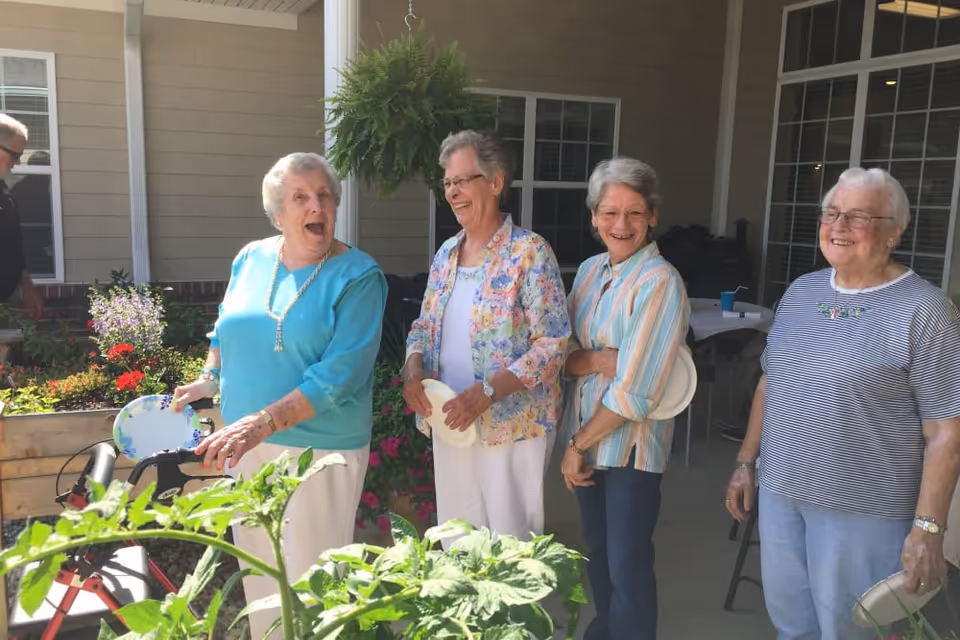 Four elderly women standing outside on a patio near a garden with plants and flowers. They are smiling and holding plates, appearing to enjoy a social gathering. The background shows windows and part of a building exterior.